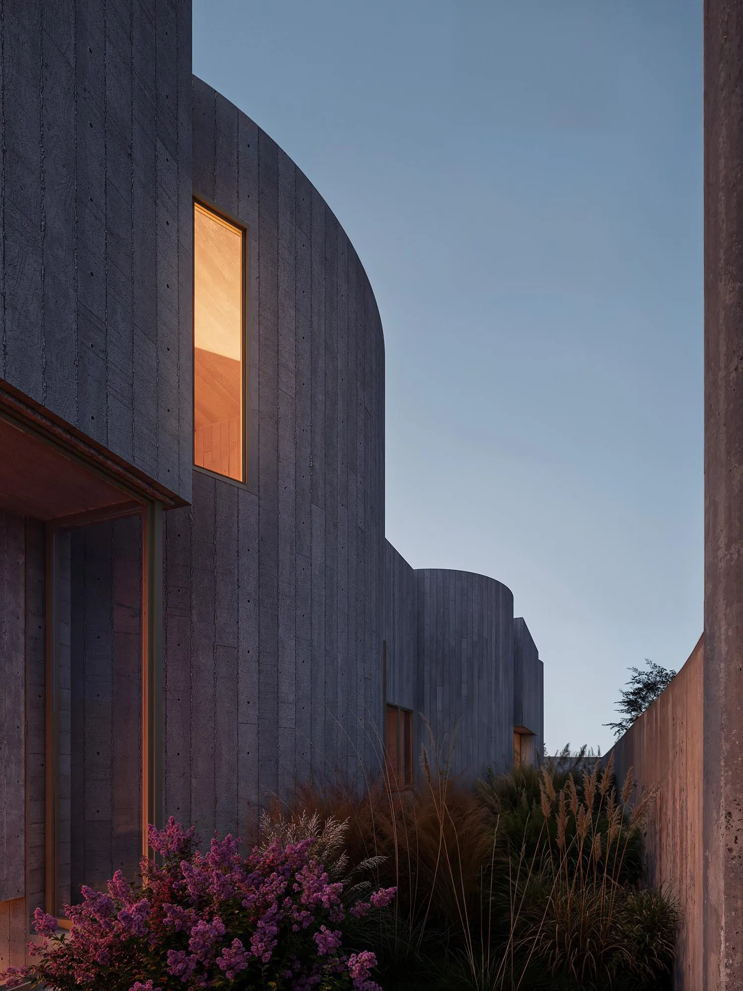 Curved facade with a lit window at dusk, pink flowers